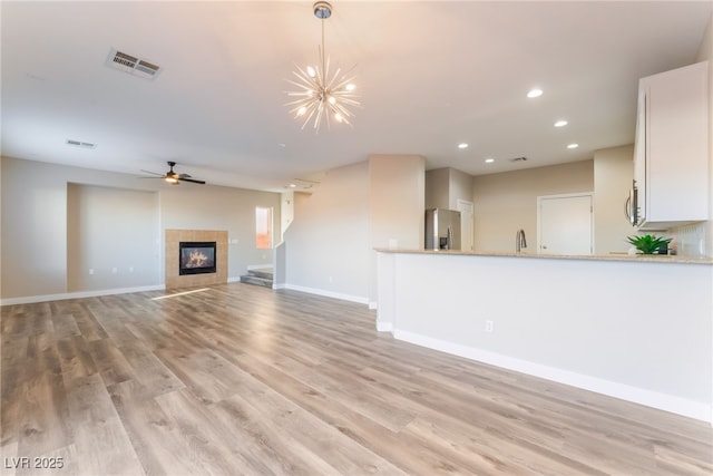 unfurnished living room with a tiled fireplace, sink, ceiling fan with notable chandelier, and light hardwood / wood-style flooring