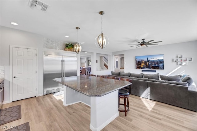 kitchen featuring a breakfast bar, dark stone counters, hanging light fixtures, stainless steel built in fridge, and light hardwood / wood-style flooring