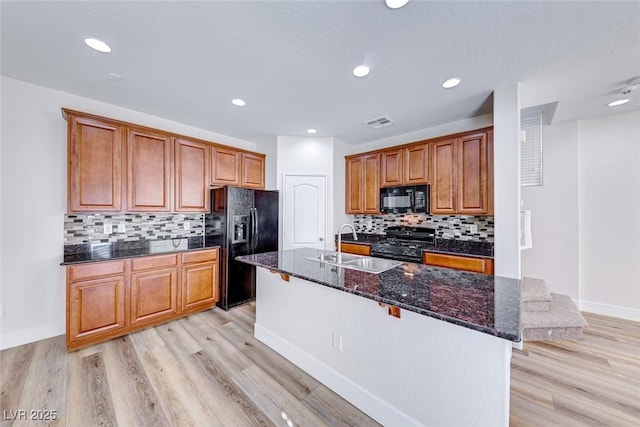 kitchen featuring sink, dark stone countertops, light hardwood / wood-style floors, black appliances, and a center island with sink