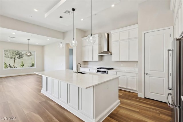 kitchen featuring white cabinetry, hanging light fixtures, a kitchen island with sink, stainless steel appliances, and wall chimney range hood