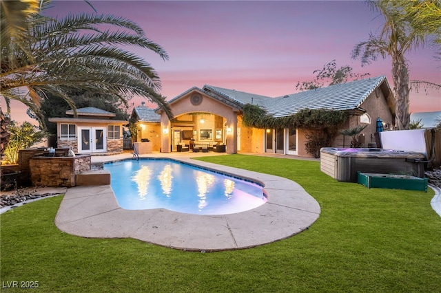 pool at dusk with french doors, a hot tub, a lawn, an outbuilding, and a patio