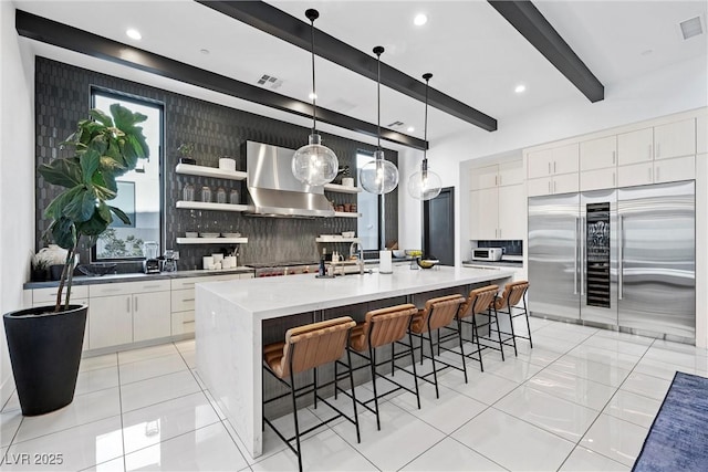 kitchen featuring white cabinets, decorative backsplash, high end fridge, a center island with sink, and wall chimney exhaust hood