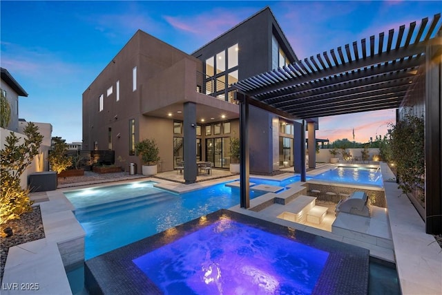 pool at dusk featuring a pergola, an outdoor kitchen, a patio area, and an in ground hot tub