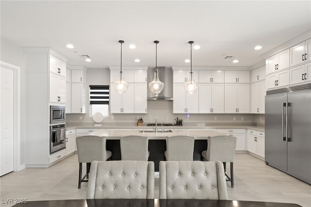 kitchen with a breakfast bar, pendant lighting, a kitchen island with sink, built in appliances, and wall chimney exhaust hood