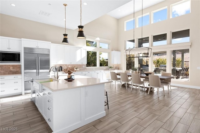 kitchen featuring a towering ceiling, white cabinets, hanging light fixtures, built in appliances, and a center island with sink