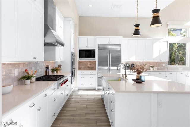 kitchen with an island with sink, pendant lighting, built in appliances, and wall chimney exhaust hood