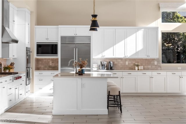 kitchen with decorative light fixtures, white cabinets, decorative backsplash, a kitchen island with sink, and built in appliances