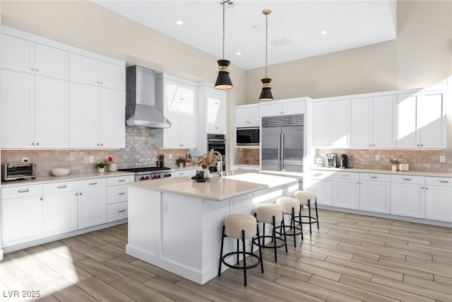 kitchen featuring built in appliances, an island with sink, white cabinets, decorative light fixtures, and wall chimney exhaust hood