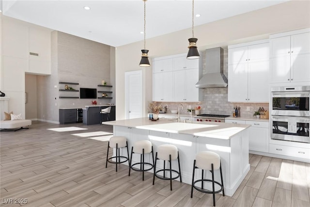 kitchen featuring appliances with stainless steel finishes, a center island with sink, a kitchen bar, decorative light fixtures, and wall chimney exhaust hood