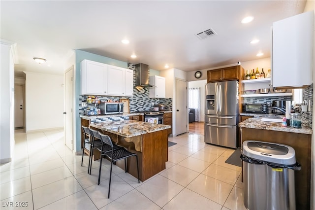 kitchen featuring wall chimney exhaust hood, white cabinetry, a kitchen island, stainless steel appliances, and light stone countertops