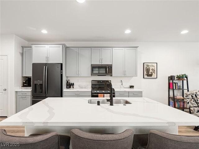 kitchen featuring sink, appliances with stainless steel finishes, gray cabinets, an island with sink, and light stone countertops