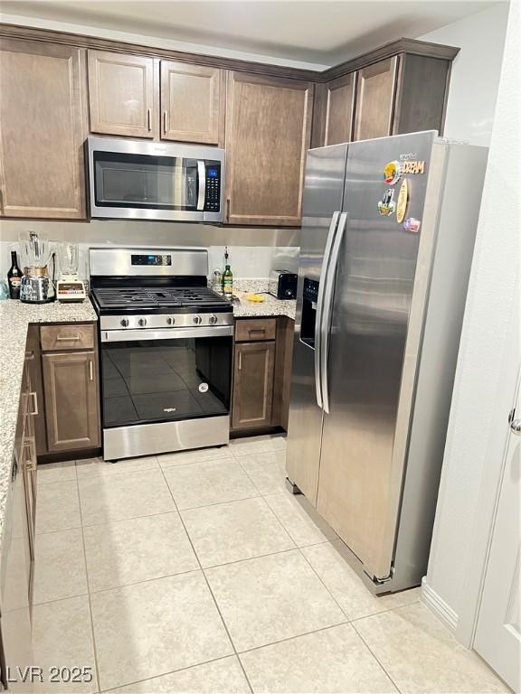 kitchen featuring dark brown cabinetry, light tile patterned floors, light stone countertops, and appliances with stainless steel finishes