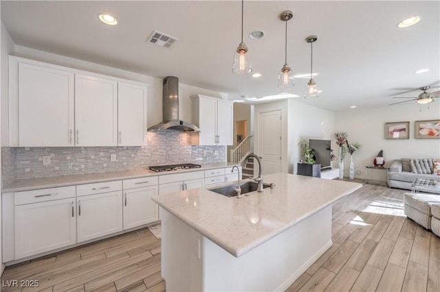 kitchen with pendant lighting, wall chimney range hood, sink, an island with sink, and white cabinets