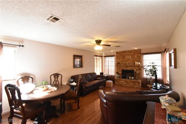 living room with a brick fireplace, dark wood-type flooring, a textured ceiling, and ceiling fan