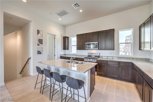 kitchen with sink, a kitchen island with sink, stainless steel appliances, light stone countertops, and a kitchen bar