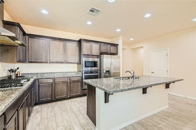 kitchen with sink, a breakfast bar, stainless steel appliances, light stone counters, and wall chimney exhaust hood