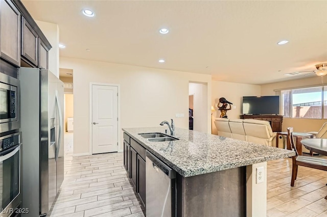 kitchen with sink, ceiling fan, a kitchen island with sink, stainless steel appliances, and light stone countertops