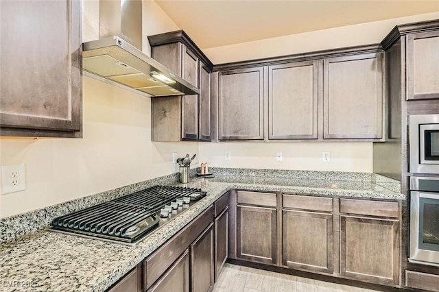 kitchen with dark brown cabinets, light stone countertops, wall chimney exhaust hood, and appliances with stainless steel finishes