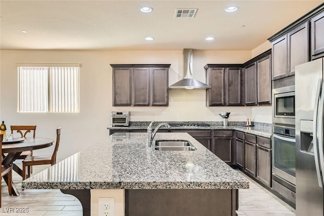 kitchen featuring wall chimney exhaust hood, appliances with stainless steel finishes, light stone countertops, and an island with sink