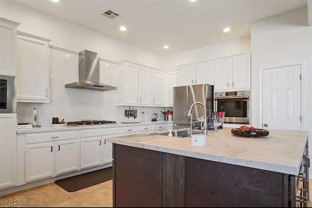 kitchen featuring wall chimney exhaust hood, light stone counters, a center island with sink, stainless steel appliances, and white cabinets