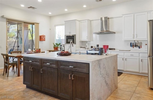 kitchen with stainless steel refrigerator, a kitchen island with sink, light stone countertops, white cabinets, and wall chimney exhaust hood