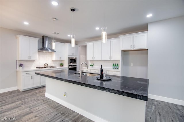 kitchen with white cabinetry, decorative light fixtures, stainless steel appliances, a kitchen island with sink, and wall chimney range hood