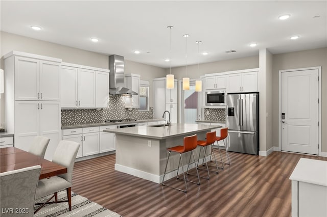 kitchen with wall chimney exhaust hood, decorative light fixtures, appliances with stainless steel finishes, a kitchen island with sink, and white cabinets