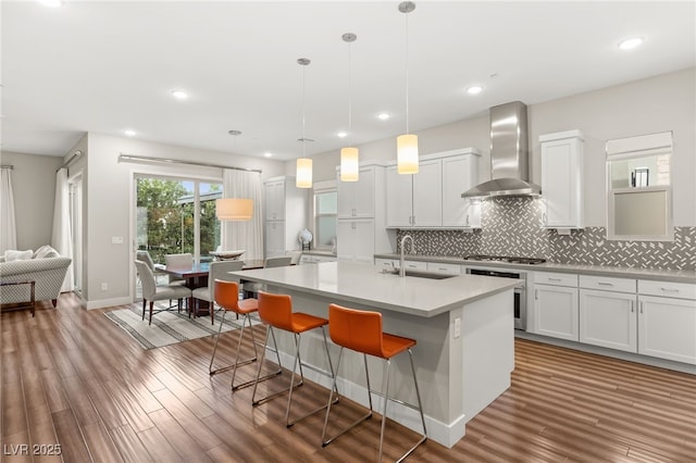 kitchen with sink, a kitchen island with sink, hanging light fixtures, white cabinets, and wall chimney exhaust hood