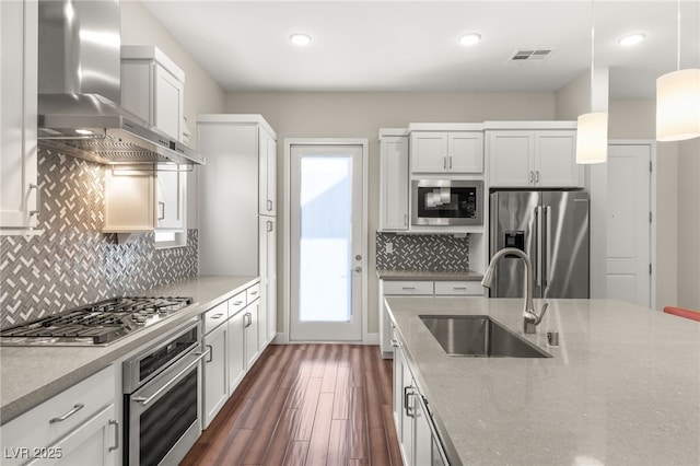 kitchen with pendant lighting, sink, white cabinets, stainless steel appliances, and wall chimney range hood