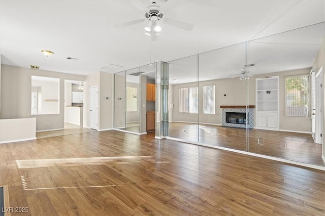 unfurnished living room with ceiling fan, a tiled fireplace, hardwood / wood-style floors, and built in features