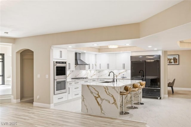 kitchen featuring white cabinetry, wall chimney range hood, stainless steel appliances, and an island with sink