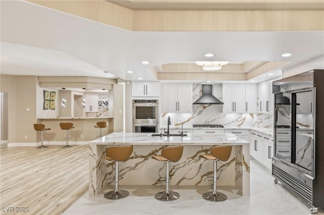 kitchen with refrigerator, white cabinets, light stone countertops, stainless steel double oven, and wall chimney exhaust hood