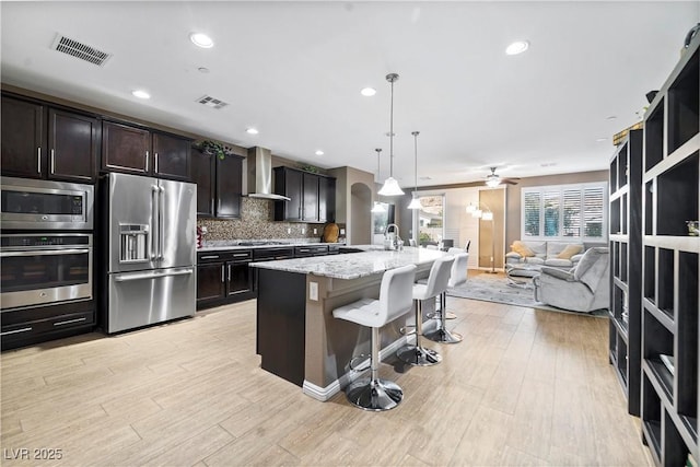 kitchen featuring pendant lighting, wall chimney range hood, stainless steel appliances, light stone countertops, and a center island with sink