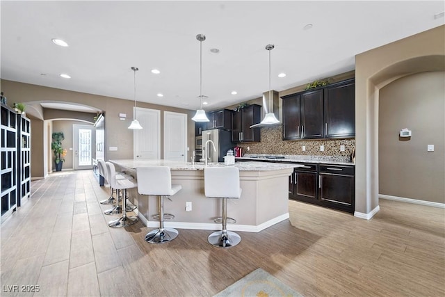 kitchen with a breakfast bar, decorative light fixtures, backsplash, a kitchen island with sink, and wall chimney exhaust hood