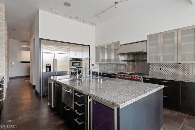 kitchen with wine cooler, wall chimney exhaust hood, an island with sink, stainless steel appliances, and backsplash