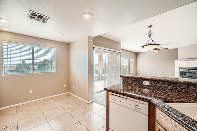 kitchen with light tile patterned flooring, dark stone countertops, white dishwasher, a tile fireplace, and pendant lighting