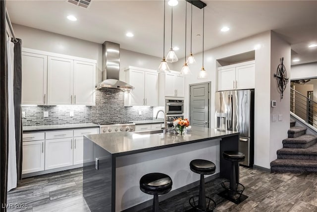 kitchen with wall chimney range hood, appliances with stainless steel finishes, white cabinetry, tasteful backsplash, and an island with sink