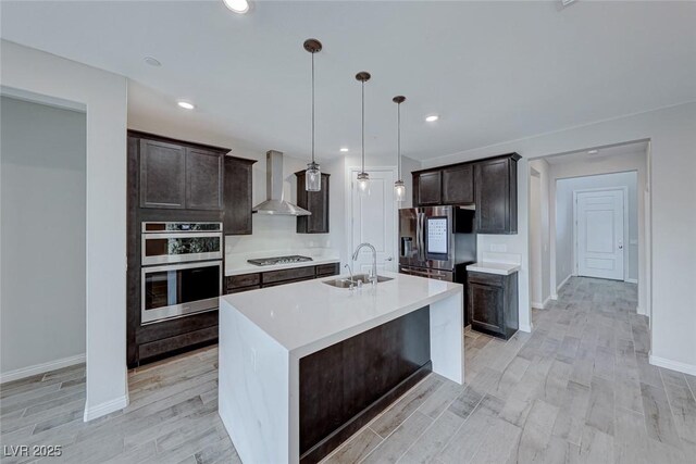 kitchen featuring pendant lighting, wall chimney range hood, sink, appliances with stainless steel finishes, and a kitchen island with sink