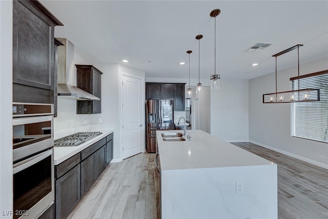 kitchen with pendant lighting, an island with sink, sink, stainless steel appliances, and wall chimney exhaust hood