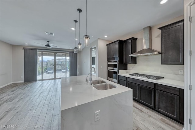 kitchen featuring sink, pendant lighting, stainless steel appliances, a kitchen island with sink, and wall chimney range hood