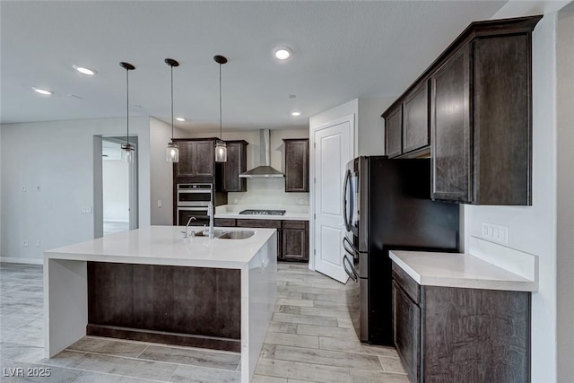 kitchen featuring wall chimney exhaust hood, sink, appliances with stainless steel finishes, pendant lighting, and a kitchen island with sink