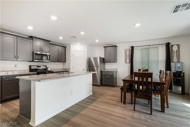 kitchen with an island with sink, appliances with stainless steel finishes, light stone counters, and light hardwood / wood-style floors