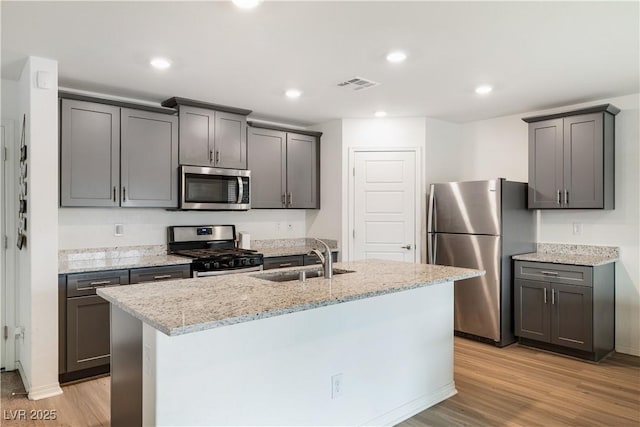 kitchen featuring stainless steel appliances, light stone countertops, a kitchen island with sink, and light hardwood / wood-style flooring