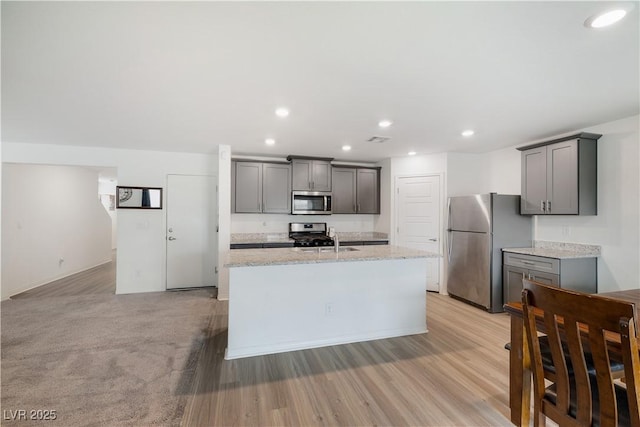 kitchen with sink, gray cabinets, appliances with stainless steel finishes, a kitchen island with sink, and light stone counters