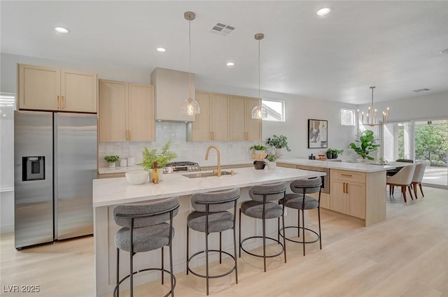 kitchen with sink, a kitchen island with sink, stainless steel refrigerator with ice dispenser, wall chimney range hood, and light wood-type flooring