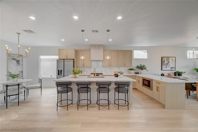 kitchen with light brown cabinetry, pendant lighting, stainless steel fridge, and a center island with sink