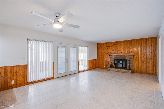 unfurnished living room with french doors, wooden walls, a stone fireplace, and ceiling fan