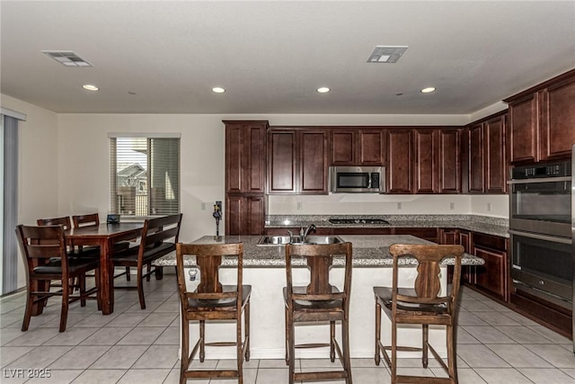 kitchen featuring sink, light tile patterned floors, appliances with stainless steel finishes, light stone countertops, and an island with sink