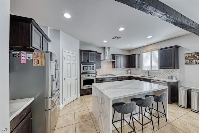 kitchen with wall chimney range hood, a breakfast bar area, stainless steel appliances, light stone countertops, and a kitchen island