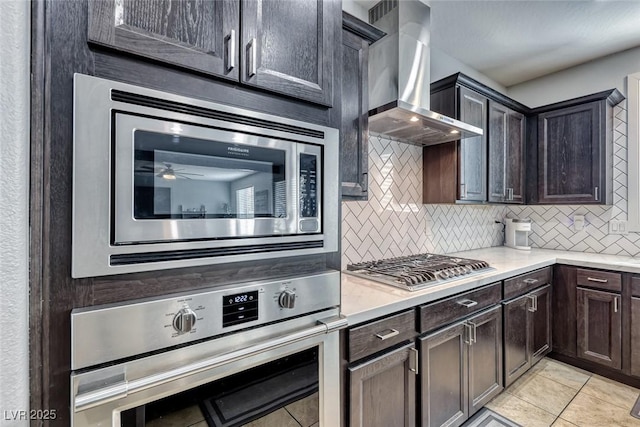 kitchen with light tile patterned floors, stainless steel appliances, dark brown cabinetry, decorative backsplash, and wall chimney exhaust hood